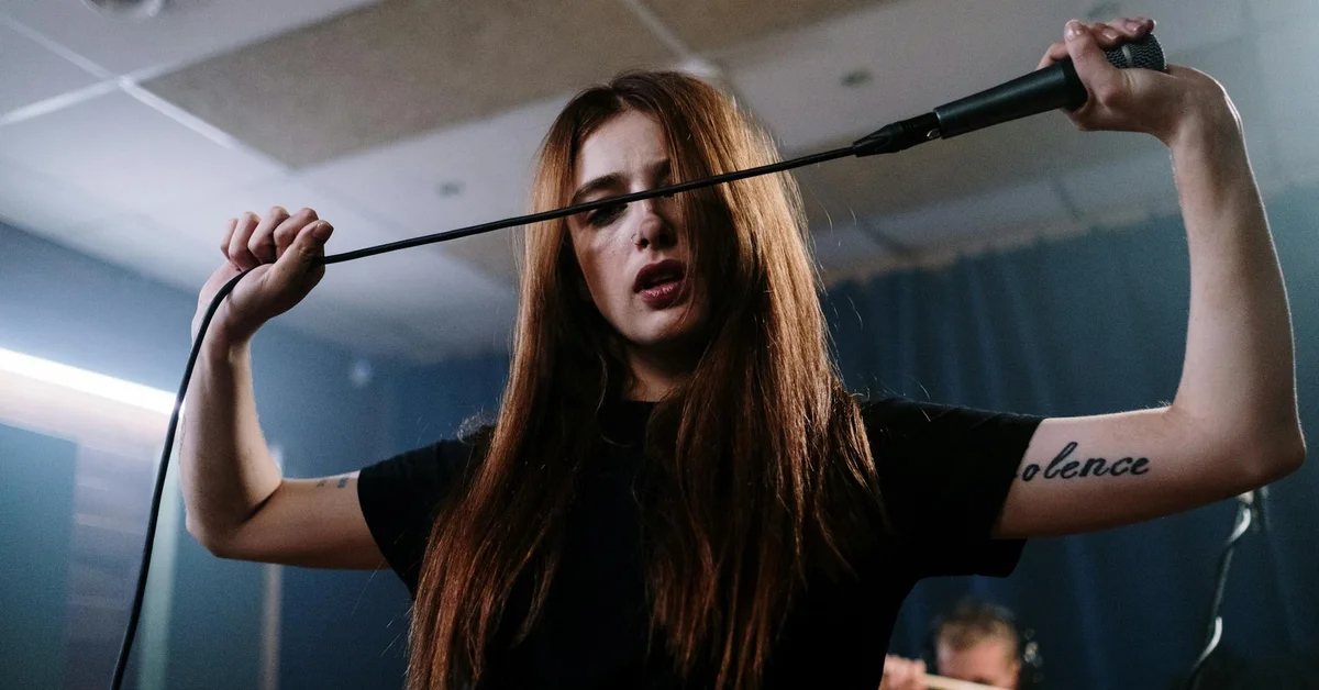 Young woman with long brown hair singing into a microphone in a studio.