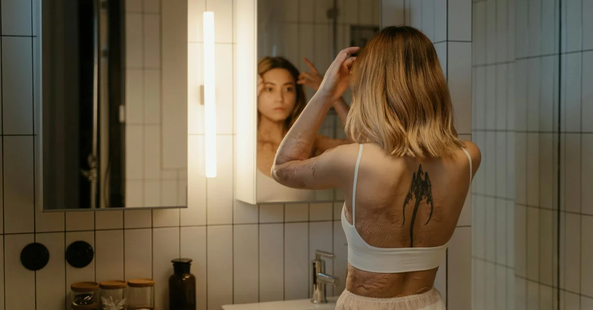 A person with a back tattoo stands in a bathroom, looking at their reflection in the mirror.