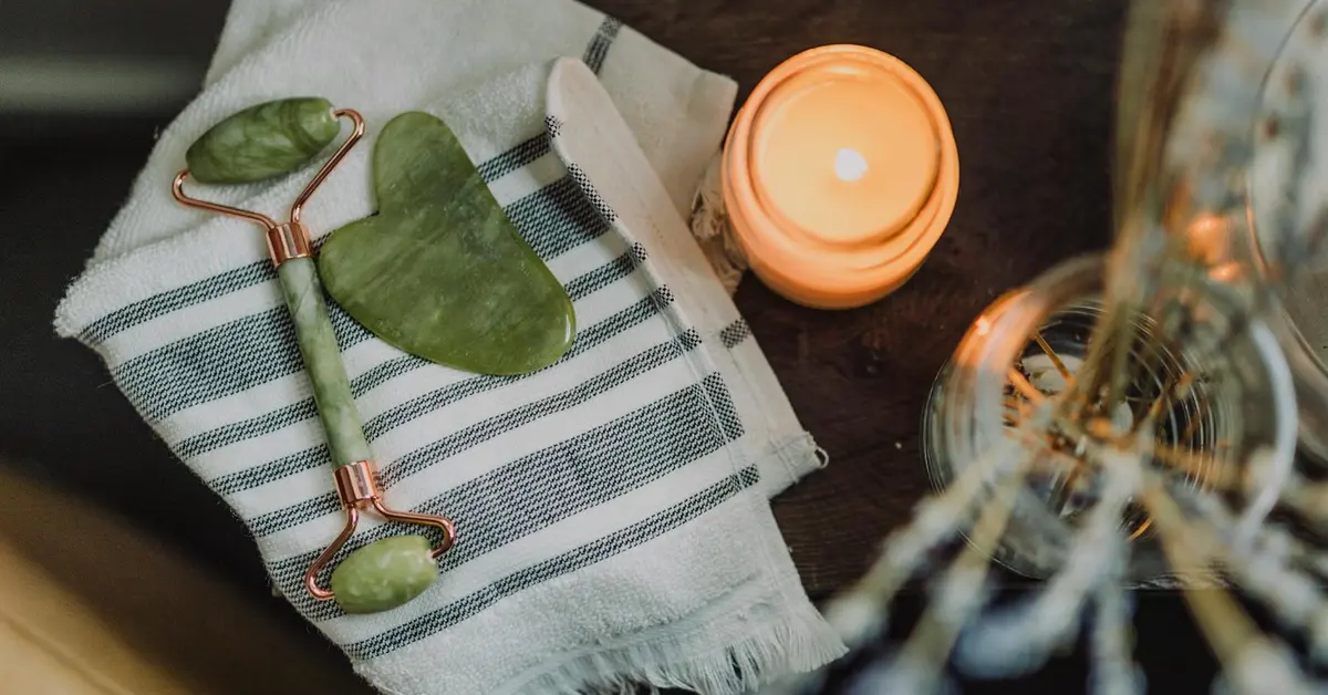 Jade roller and skincare tools placed on a striped cloth with a lit candle in a soft-lit spa-like setting.