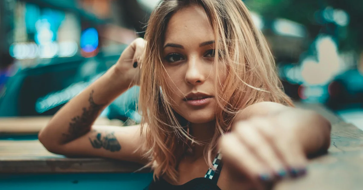 Portrait of a tattooed person leaning on a surface with a blurred urban background.
