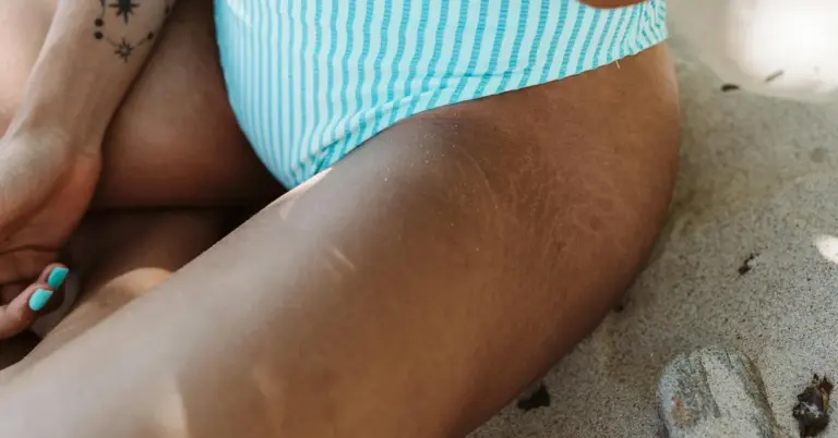 Close-up of a person on a sandy beach wearing a blue striped swimsuit, with a small tattoo visible on the upper arm.