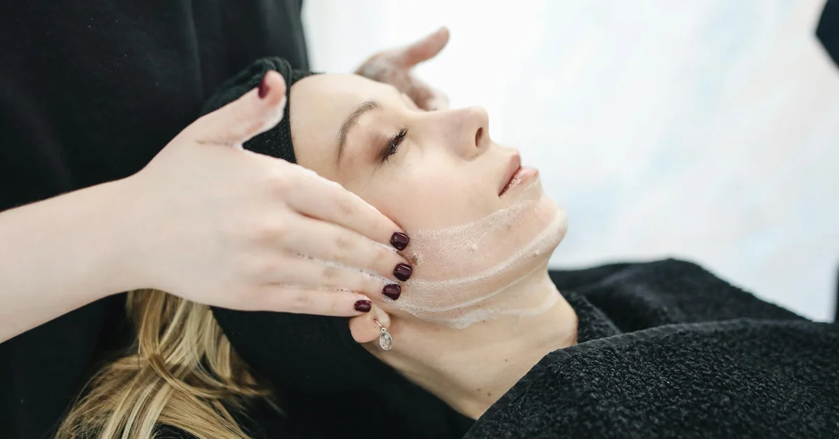 Close-up of a client with eyes closed as a technician applies topical treatment to the face, illustrating professional skincare safety steps relevant to numbing creams used in tattoo removal.