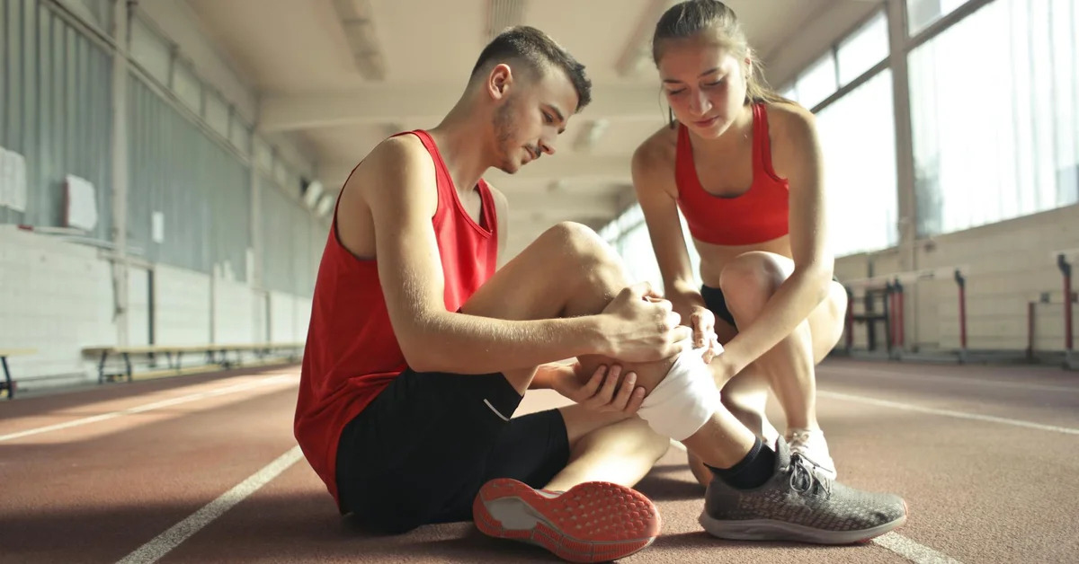 Two people on an indoor track; one person sits with an ankle injury while the other tends to it, illustrating care and discomfort.