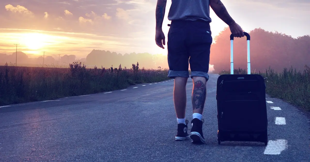 Athlete or traveler in shorts and sneakers walking along a road at sunset, pulling a rolling suitcase; a calf tattoo is visible.