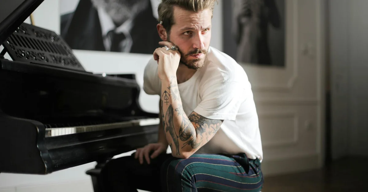 A tattooed man sits beside a grand piano in a bright, elegant room, looking thoughtful.