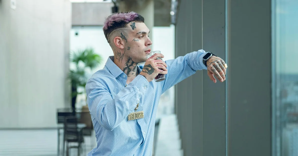 Person with multiple facial and neck tattoos wearing a light blue shirt looking out a window in a modern clinical setting.