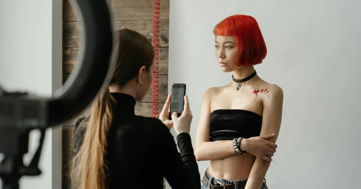 Model with a bright red bob hairstyle in a black strapless top, displaying a small shoulder tattoo while a photographer takes a picture.