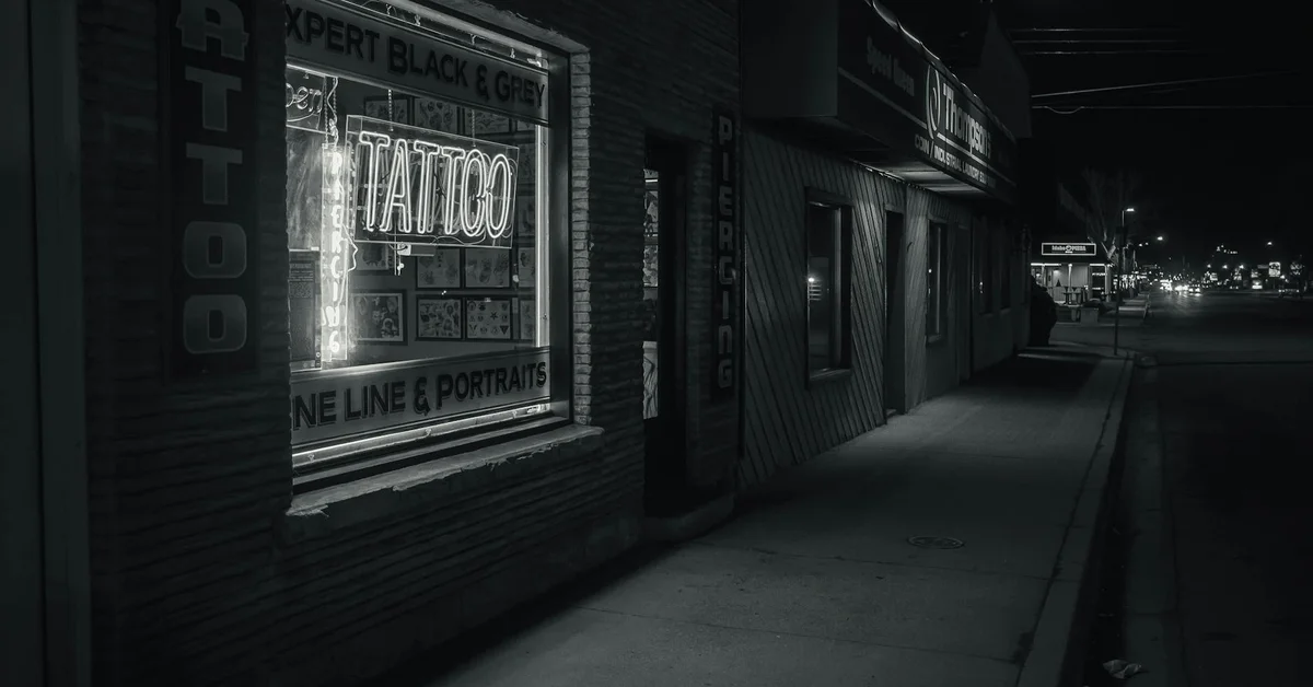 Night-time street view of a tattoo studio with a bright neon 'TATTOO' sign in the window
