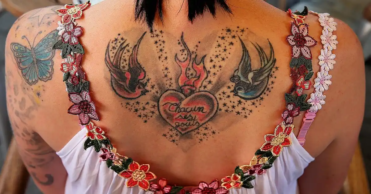 Back view of a person wearing a floral dress, showing a large back tattoo with a central heart and script between two birds, surrounded by dotted shading and decorative floral elements along the shoulders.