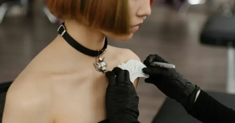Close-up of a professional wearing gloves removing a white bandage from a fresh tattoo on a client's shoulder.