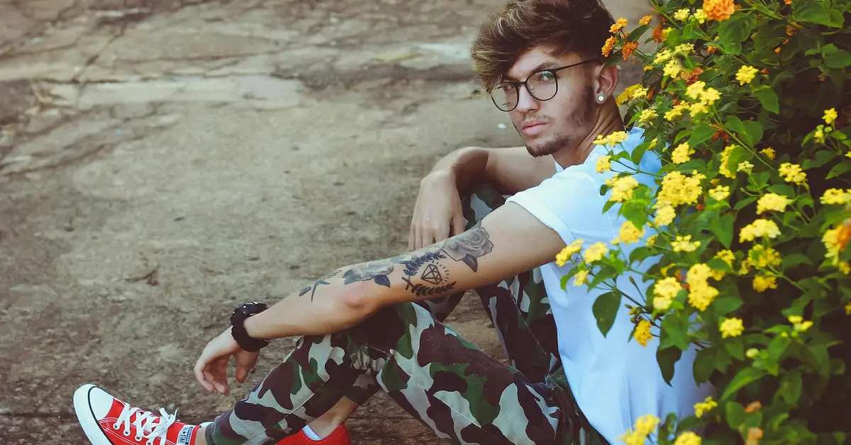 Young person with multiple tattoos sitting outdoors near yellow flowers, wearing glasses, a white t-shirt, camouflage pants, and red sneakers.