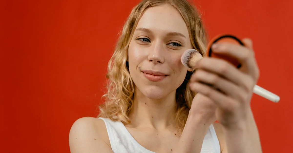 A young woman with wavy blonde hair applies makeup with a brush in front of a red background.