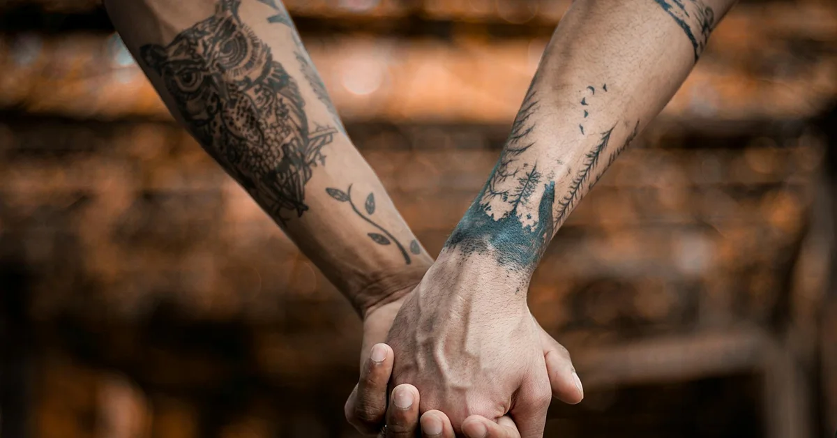 Two tattooed forearms crossed and hands clasped, photographed with a warm, blurred background.