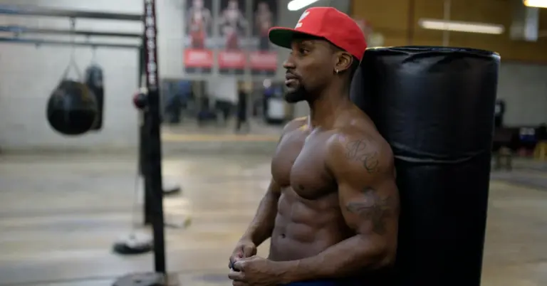 A shirtless, tattooed man wearing a red cap sits by a large punching bag in a gym.