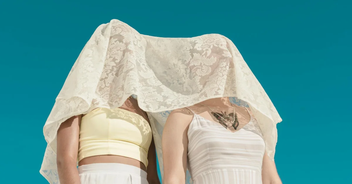 Two people standing side by side with a sheer lace fabric draped over their heads, revealing their light-colored tops.