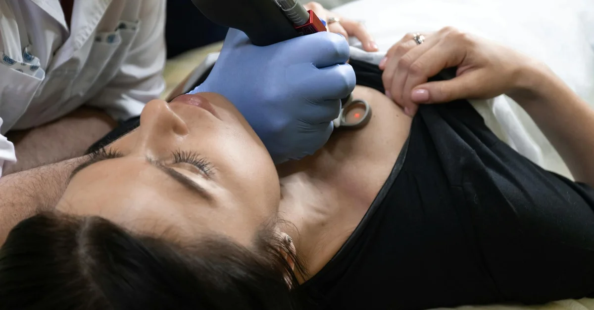 A medical professional in blue gloves uses a handheld laser device on a patient's neck and upper chest during a tattoo removal treatment.