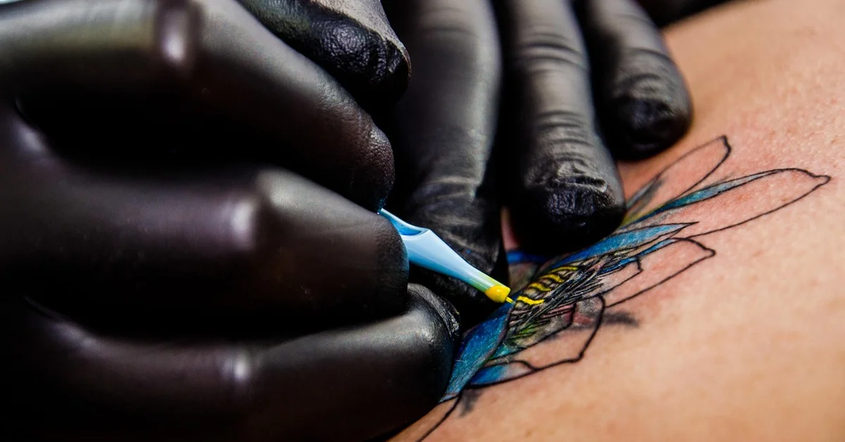 Close-up of a gloved hand outlining a colorful tattoo on skin with a blue marker tool.