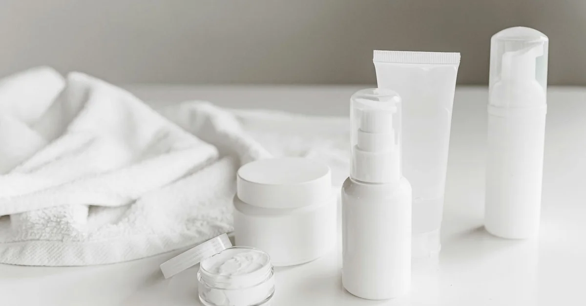 A collection of white skincare product containers—bottles, tubes, and jars—displayed on a white surface with a folded towel in the background.