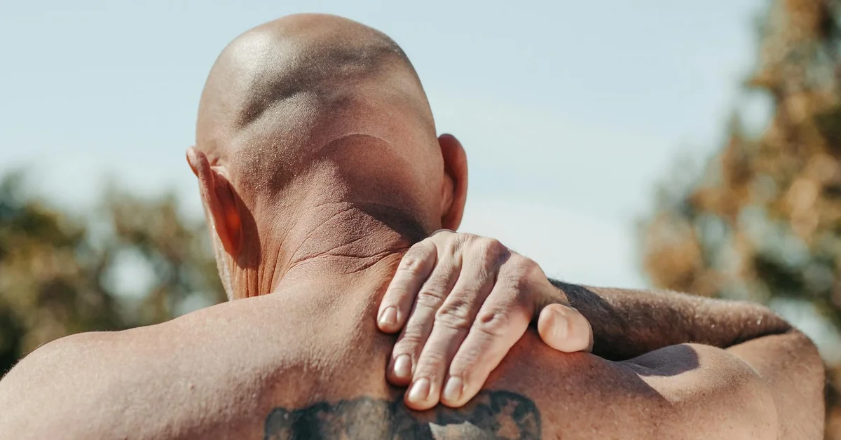 Outdoor back view of a shirtless man with a tattoo on his upper back, his hands resting on his shoulders.