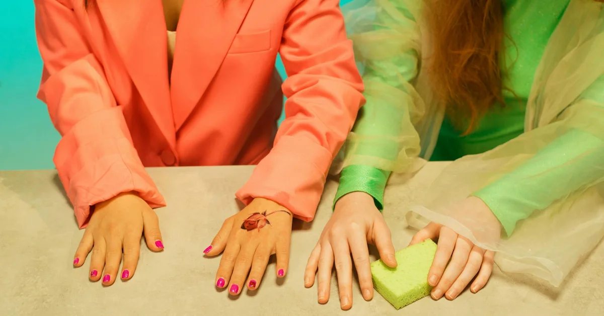 Two hands resting on a table; one hand has bright pink nail polish and a small tattoo, the other shows a green-sleeved arm nearby.