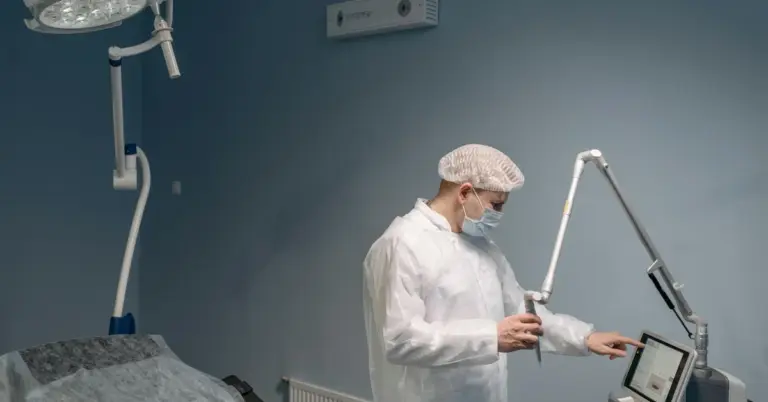 Medical professional in a sterile gown, mask, and cap operating a laser device in a clinical setting.