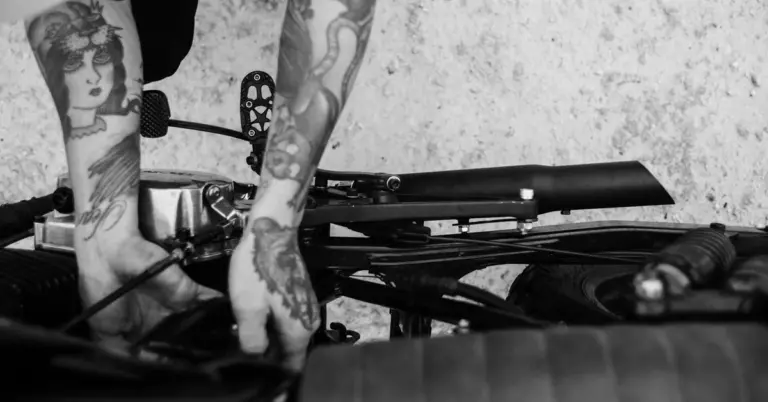 Black-and-white close-up of tattooed forearms beside dark mechanical equipment
