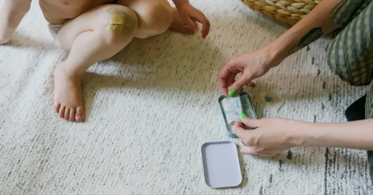 A caregiver applies a small adhesive patch to a baby's leg on a light-colored rug.