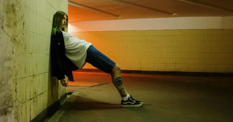Person leaning against a wall in a dimly lit underground parking area, with a visible calf tattoo.