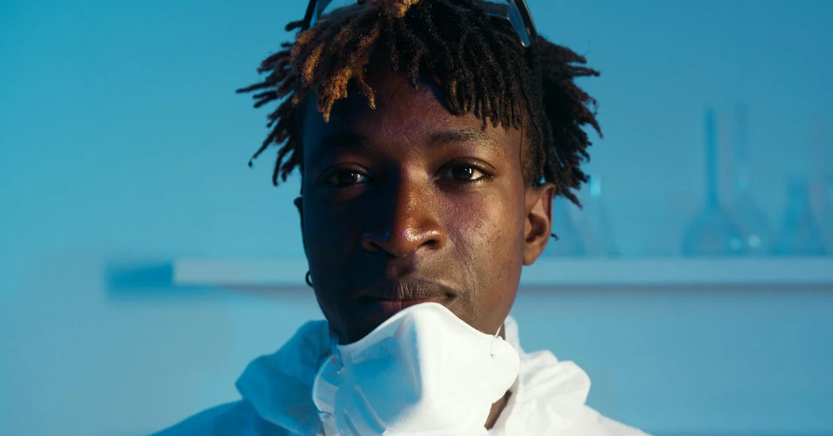 Portrait of a person in a clinical setting with blue lighting, wearing protective gear and a mask around the chin.