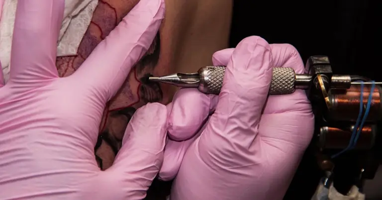 Close-up of a practitioner wearing pink gloves using a tattoo removal tool on a client’s skin, focusing on the area where scabs may form.