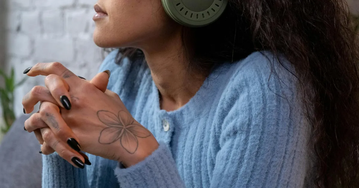 Close-up side profile of a person wearing a blue knit sweater and large headphones, hands clasped, with a flower tattoo on the back of the hand.