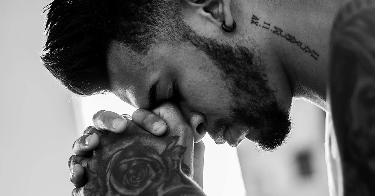 Black-and-white portrait of a man with neck and hand tattoos, head bowed and hands clasped near his face.