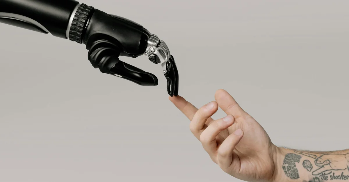 Close-up of a black robotic hand reaching toward a tattooed human finger against a neutral gray background, symbolizing the choice between removal and cover-up.