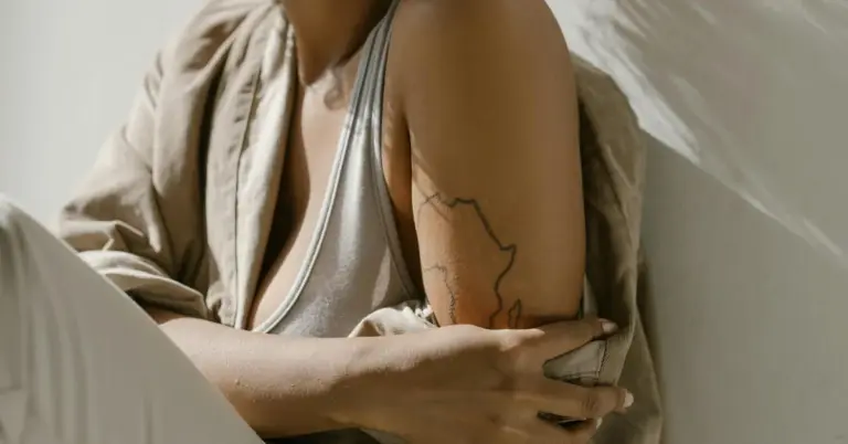 Close-up of a person’s upper arm and shoulder with a simple line tattoo along the arm; the person wears a light beige jacket and a white top.