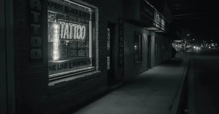 Black-and-white street view at night showing a tattoo shop storefront with a glowing neon sign in the window