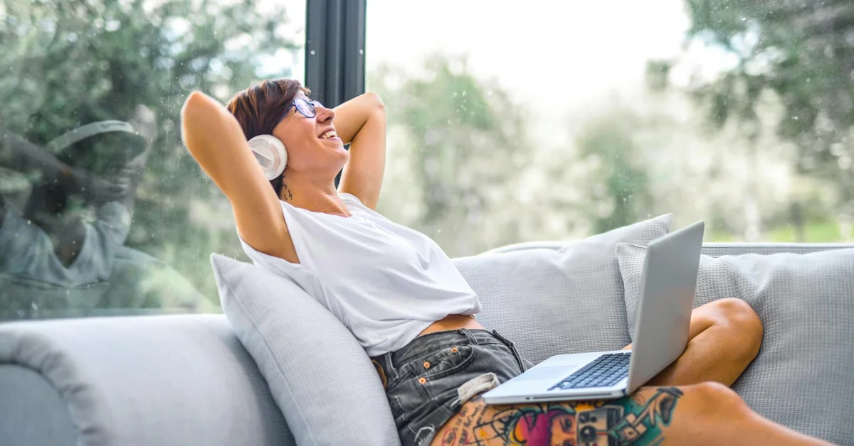 Person lounging on a sofa with headphones and a laptop, exposing colorful leg tattoos.