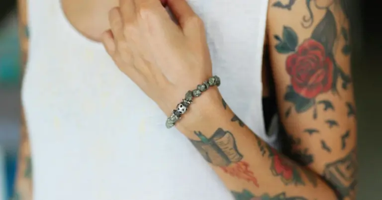 Close-up of a tattooed forearm with a red rose and other designs, wearing a white sleeveless shirt and a beaded bracelet.