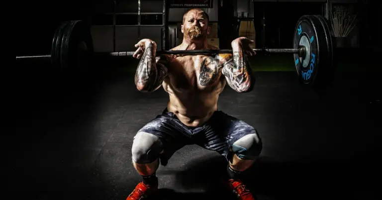 Tattooed man performing a barbell squat in a dimly lit gym.