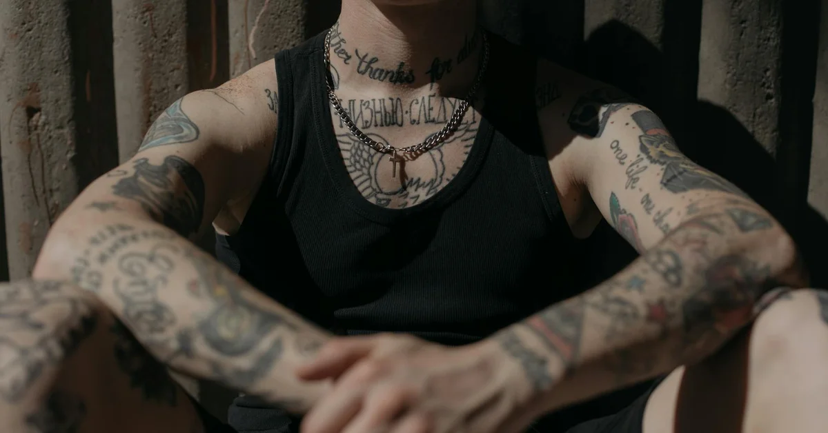 Close-up of tattooed forearms and chest of a person wearing a black tank top, sitting with arms crossed.