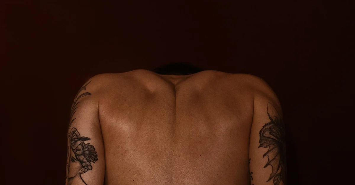 Back view of a person with tattoos on the upper arms and shoulders, shown in dim lighting.