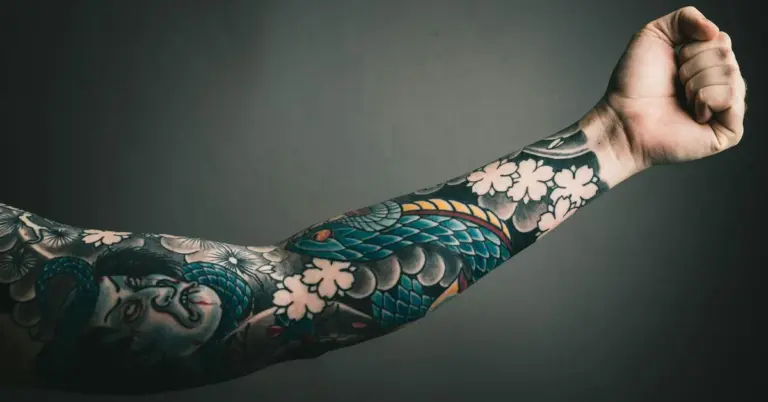 Close-up of a forearm covered in a colorful tattoo sleeve featuring a blue-green dragon and white flowers against a dark background.