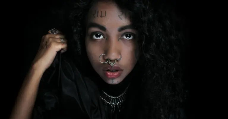 Close-up portrait of a person with facial tattoos and a nose ring against a dark background.