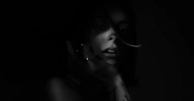 Close-up portrait of a person in low lighting, with their hand near their face in a moody, high-contrast shot.