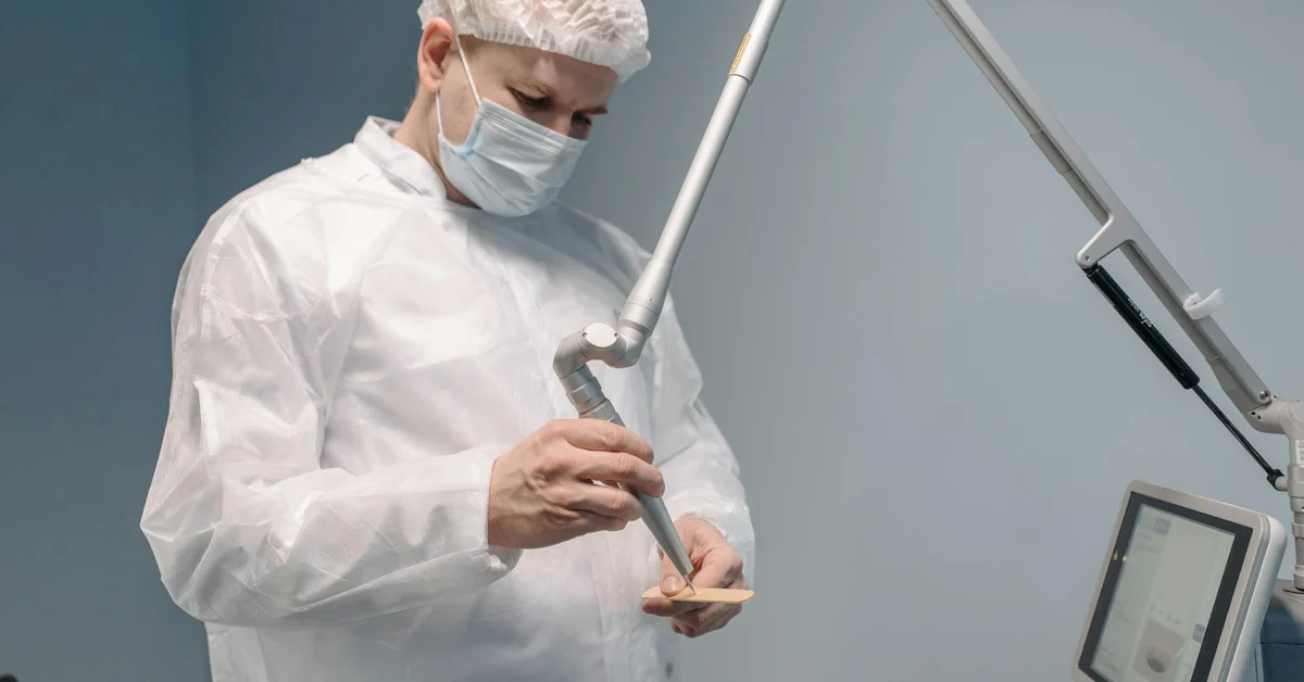 Medical professional in sterile PPE adjusts a dental instrument beside a monitor.