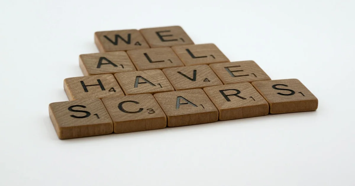 Scrabble tiles spelling 'WE ALL HAVE SCARS' arranged on a white background