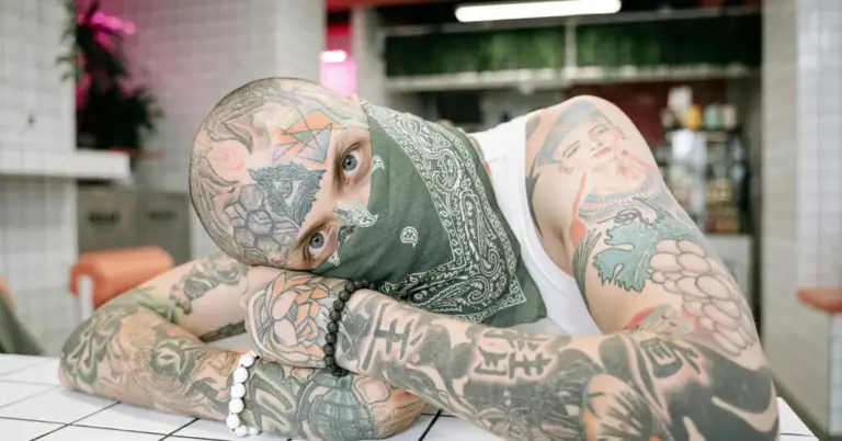 A heavily tattooed person with a bandana covering the lower face, leaning on a tiled counter in a casual setting.