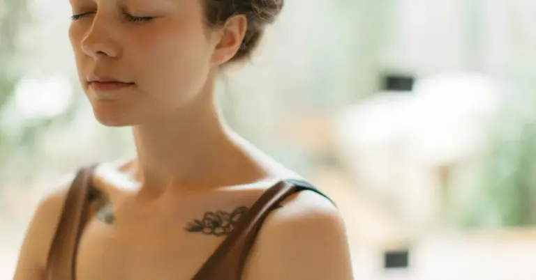 Close-up of a woman with small tattoos on her shoulder and collarbone, eyes downcast, wearing a brown top.